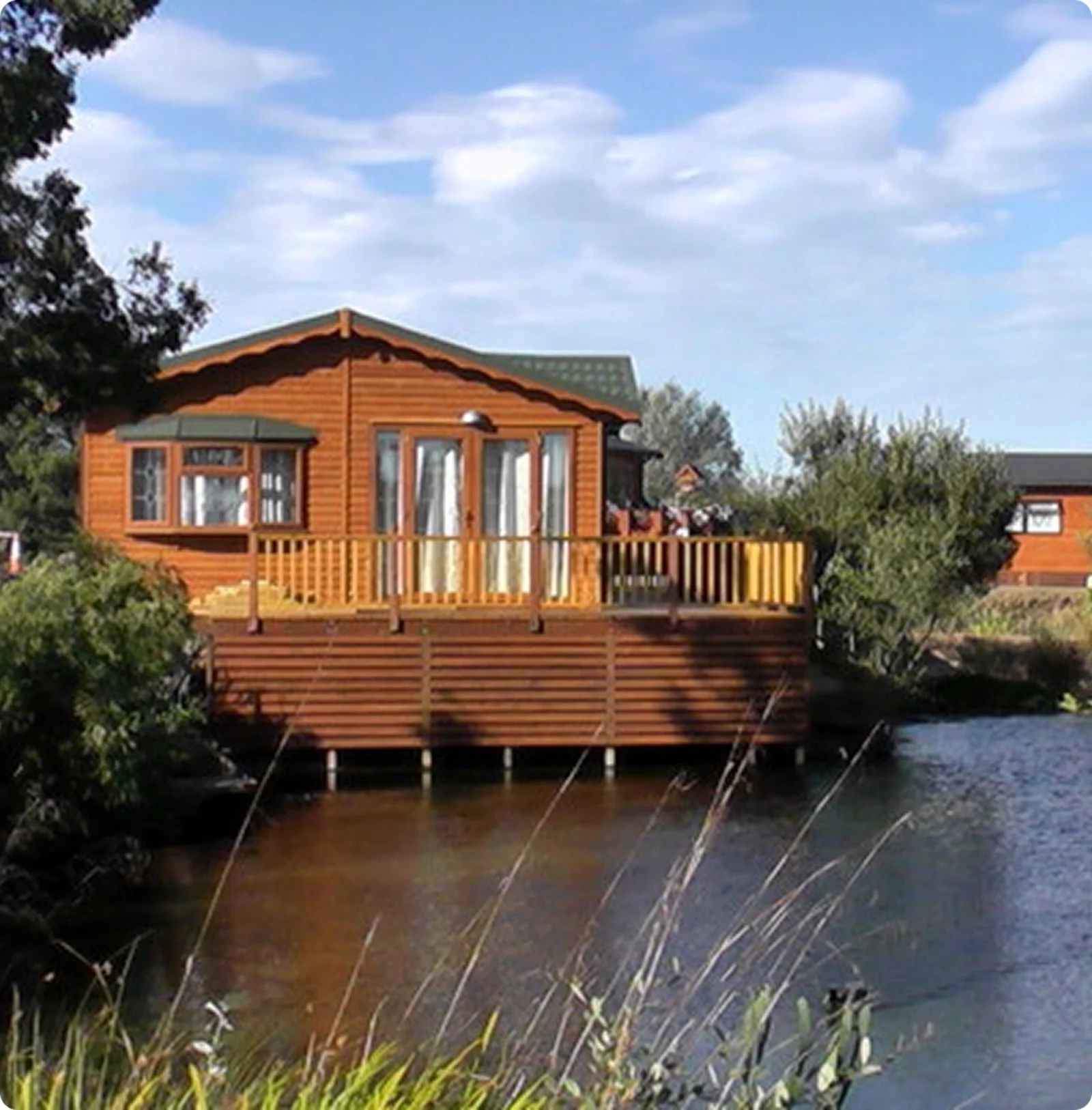 A wooden house on stilts with a deck overlooking a body of water, surrounded by trees and under a partly cloudy sky.