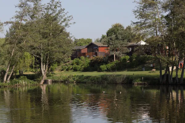 House with wooden exterior and balcony overlooking a pond, surrounded by trees and lush greenery, with ducks swimming in the water.