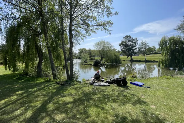 A person is sitting on a chair near a calm lake surrounded by trees, with fishing gear and a blue flotation device on the grass in a sunny park.