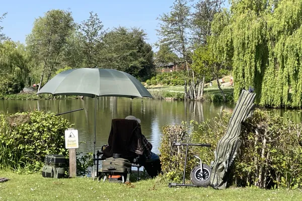 A person is sitting by a river with an umbrella, surrounded by trees and bushes, with fishing equipment and a sign that warns of danger near the water.